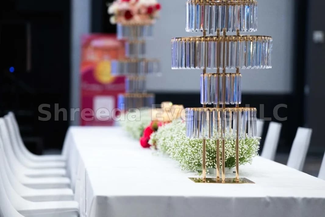 Long banquet table with a white tablecloth, white chairs and a tall crystal and floral centerpiece