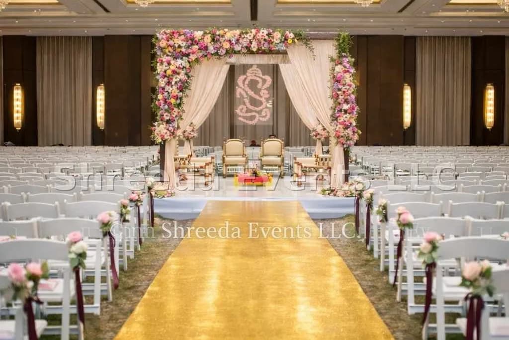 Indoor wedding ceremony with a pink and white floral mandap, white chairs and a gold carpet aisle