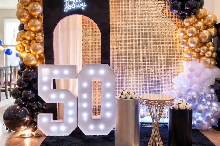 50th birthday setup with a gold shimmer wall, a black arch with a Happy Birthday sign, black and gold balloons and lit marquee numbers