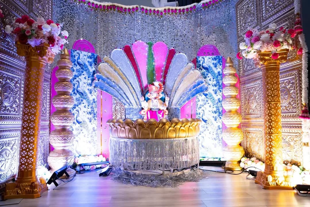 Ganesh idol on a crystal-draped lotus base before a fan of blue, purple and pink panels, with golden pillars and embossed silver walls