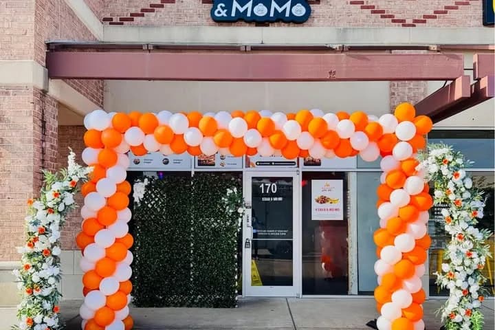 Orange and white balloon arch with greenery and white flowers around the front door of Kebab & Momo