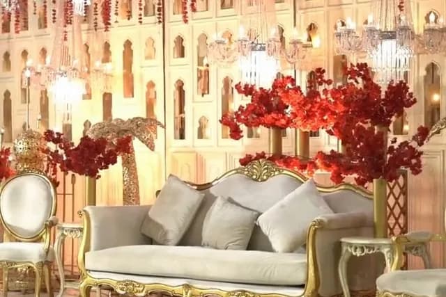 Reception stage with red blossom trees, an ivory arched panel backdrop, hanging crystals, a cream tufted sofa and gold chairs