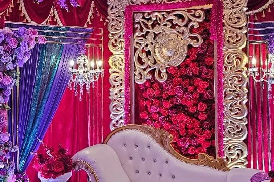 Wedding stage with a red rose wall in an ornate ivory and gold frame, hanging purple florals, jewel-toned drapes and a white chaise sofa