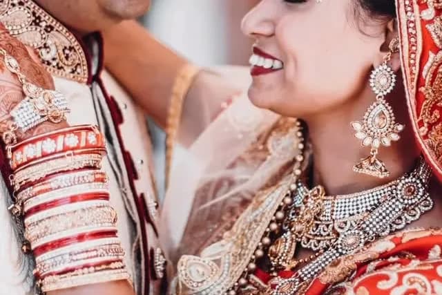 Bride in a red and gold bridal outfit with gold jewelry beside the groom in an embroidered sherwani