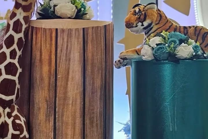 Jungle birthday setup with a giraffe figure, wood-textured plinths topped with white flowers, a teal cylinder and a tiger figure