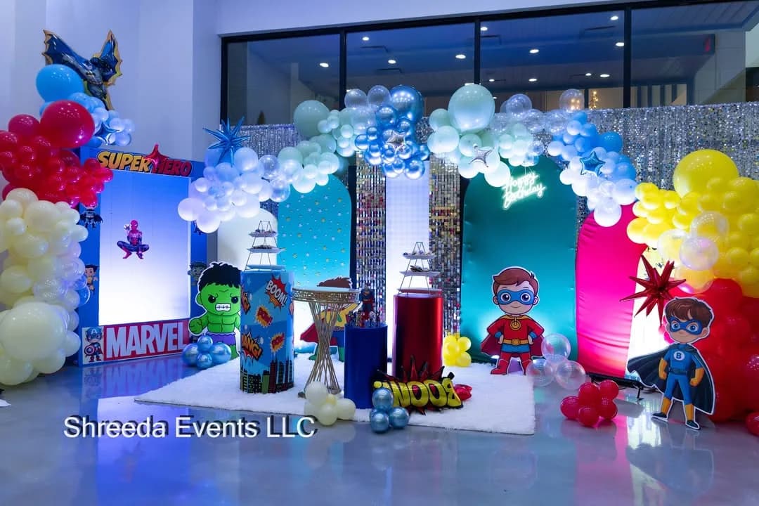 Superhero birthday backdrop with red, blue, yellow and white balloon garlands, comic panels, a teal shimmer wall and character cutouts
